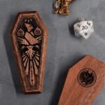 2 closed Mini Dice Sarcophagi, showing top and bottom engraving, next to D20 and miniature figurine