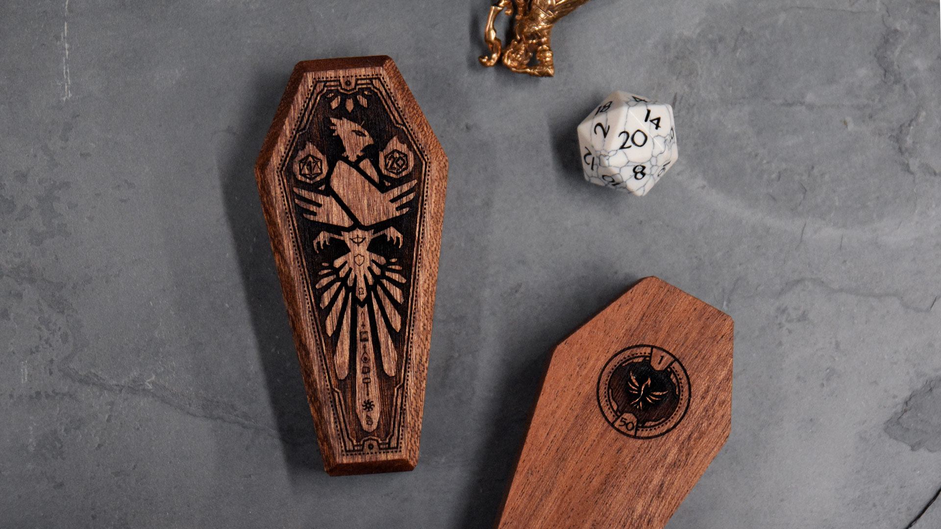 2 closed Mini Dice Sarcophagi, showing top and bottom engraving, next to D20 and miniature figurine