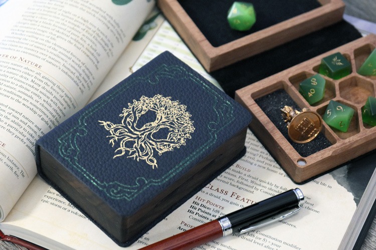 two mini spellbook dice boxes with blue leather, foil pressed with a gold tree, with a set of green cats eye dice and a Hero Forge miniature on a D&D player's manual