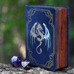 mini spellbook dice box with blue leather, foil pressed with a holographic winged dragon with two amethyst gemstone dice